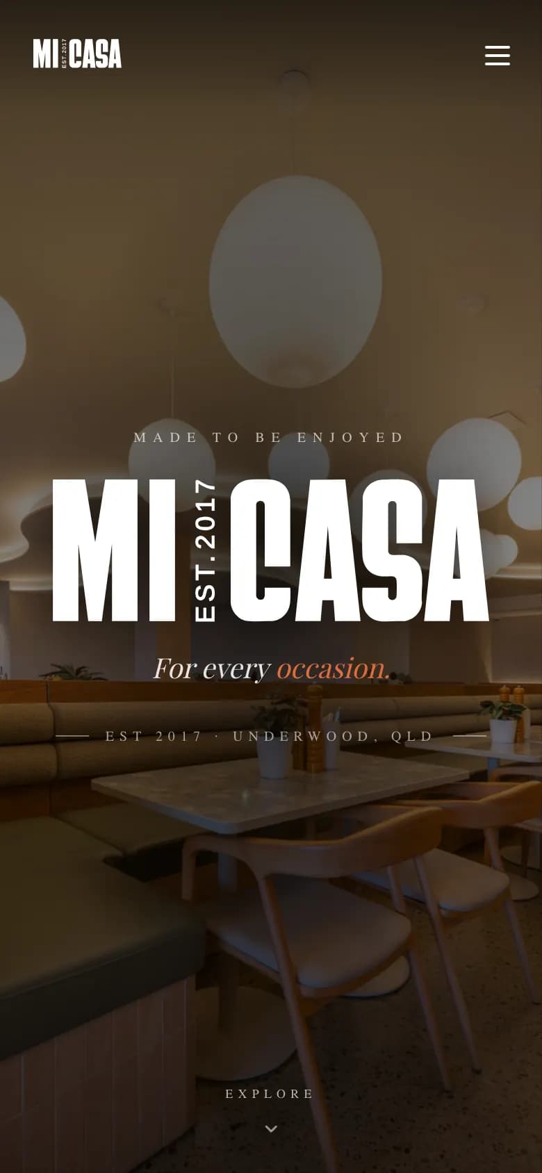 Mobile homepage hero: the MI CASA wordmark with vertical EST.2017 fills the screen width over the dimmed dining-room photo, with a menu icon top right and an Explore cue at the bottom